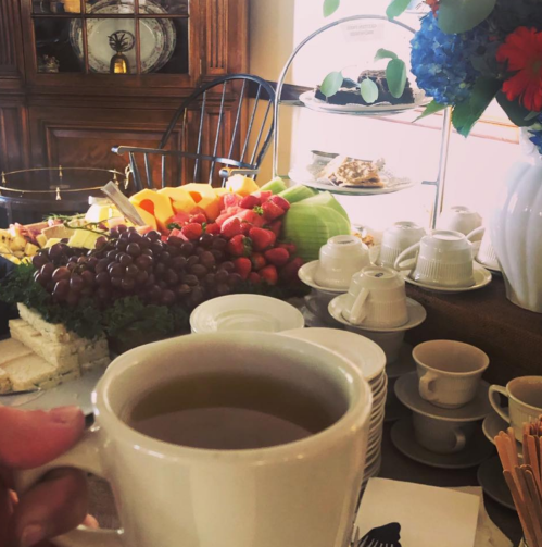 A hand holds a cup of tea in front of a table filled with fruit, pastries, and tea cups.