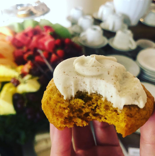 A hand holding a partially eaten cookie with frosting, surrounded by a colorful fruit platter and tea set in the background.