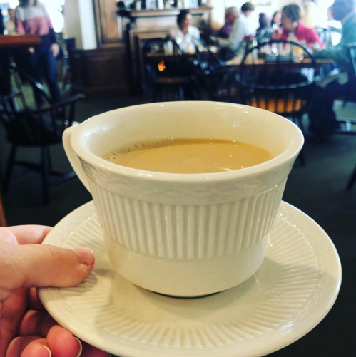 A hand holding a large white coffee cup on a saucer, with a busy café in the background.