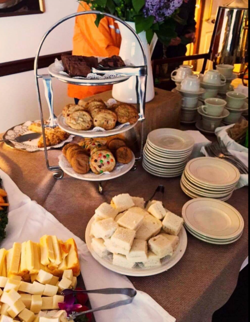 A tiered stand with cookies and brownies, surrounded by cheese, sandwiches, and plates on a table.