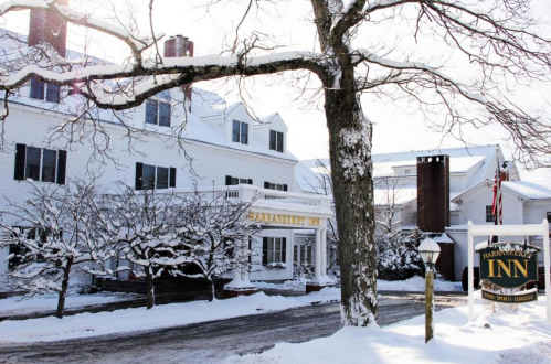 A snowy scene of the Harraseeket Inn, featuring white buildings and a sign, surrounded by bare trees.