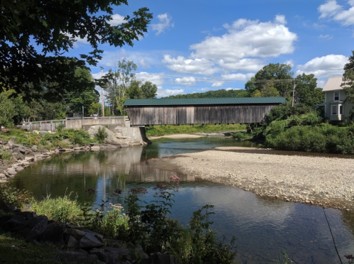 A wooden covered bridge spans a calm river, surrounded by greenery and under a blue sky with fluffy clouds.