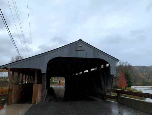 A covered bridge on a rainy day, with trees in autumn colors and a river nearby.