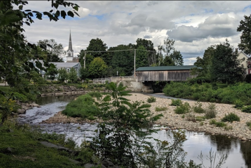 A serene river scene with a covered bridge, lush greenery, and a church steeple in the background under a cloudy sky.