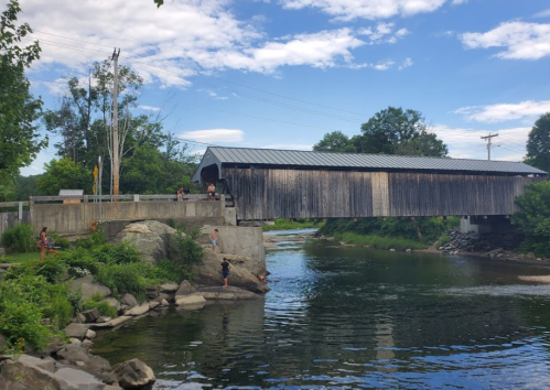 A wooden covered bridge spans a river, surrounded by greenery and people enjoying the outdoors on a sunny day.