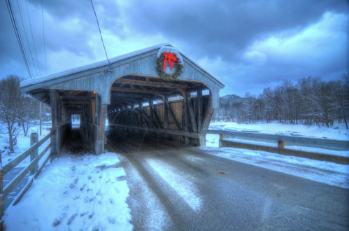 A snowy scene featuring a covered bridge adorned with a festive wreath, surrounded by winter trees and a cloudy sky.