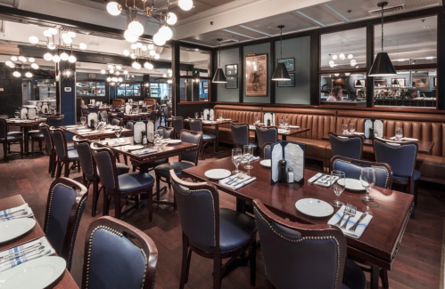 A stylish restaurant interior with wooden tables, blue chairs, and warm lighting, set for dining.