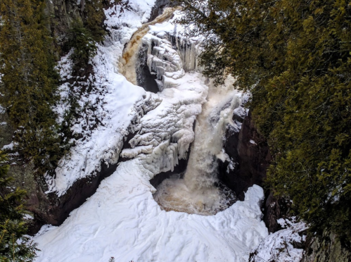 A frozen waterfall cascading over rocky cliffs, surrounded by snow and evergreen trees.