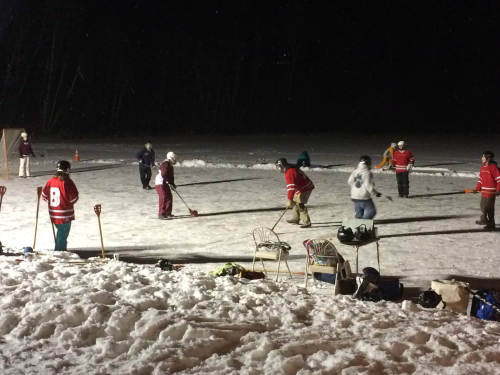 A group of people playing hockey on an outdoor rink at night, surrounded by snow and trees in the background.