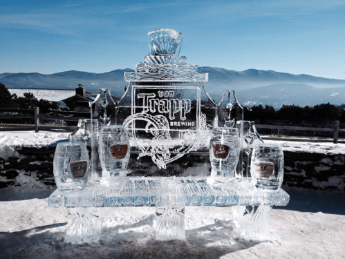 Ice sculpture of a brewery logo with barrels and bottles, set against a snowy mountain backdrop.