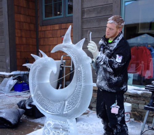 A man carves an intricate ice sculpture of a dragon-like design in a snowy outdoor setting.