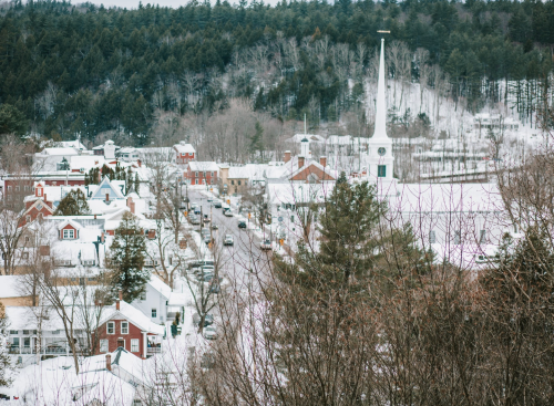 A snowy village scene with quaint houses, a church steeple, and trees in the background, creating a serene winter landscape.
