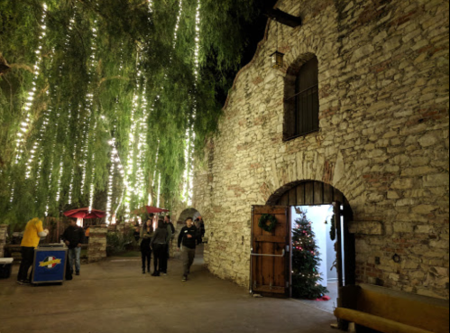 A stone building with a wooden door, surrounded by trees adorned with lights, and people walking nearby.