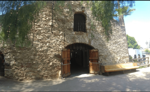 Historic stone building with wooden doors and a bench outside, surrounded by greenery and palm trees.