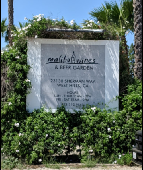 Sign for Malibu Wines & Beer Garden, located at 23130 Sherman Way, West Hills, CA, with hours listed. Surrounded by greenery.