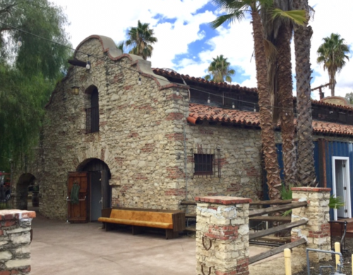 Historic stone building with a tiled roof, surrounded by palm trees and a rustic wooden bench in front.