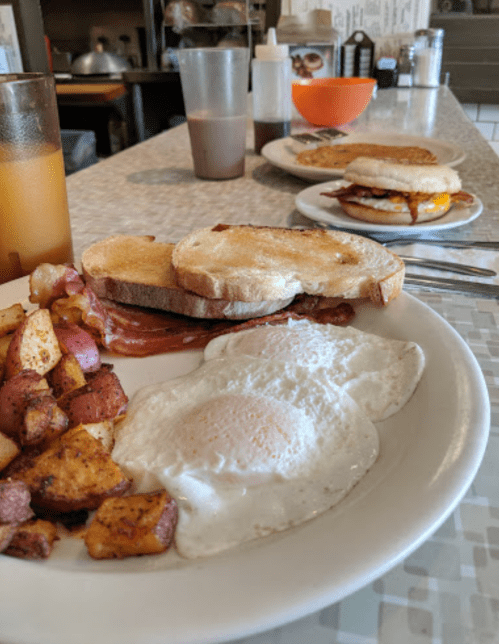 A plate with two fried eggs, toast, bacon, and seasoned potatoes, with drinks and another dish in the background.
