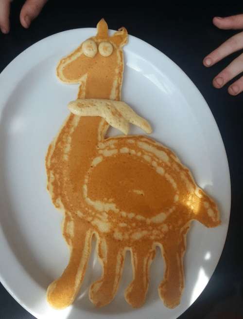 A creatively shaped pancake resembling a giraffe, served on a white plate with hands in the background.
