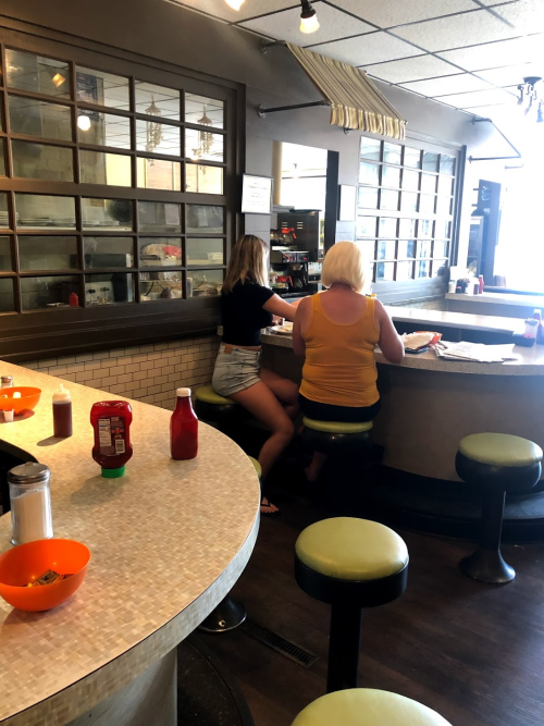 Two women sit at a diner counter, one in a black top and shorts, the other in a yellow top, with condiments on the table.