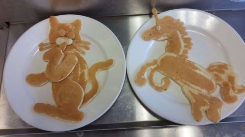 Two plates of pancakes shaped like a cat and a horse, creatively arranged with details for eyes and features.