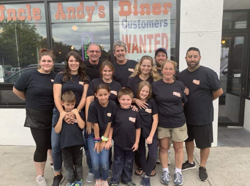 A group of 14 people in matching black shirts poses outside Uncle Andy's Diner, smiling in front of a "Customers Wanted" sign.