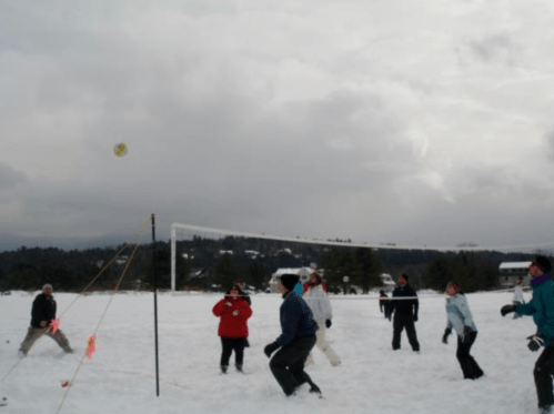 A group of people playing volleyball in the snow, with a cloudy sky and trees in the background.