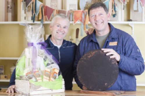 Two smiling men stand together, one holding a round wooden board and the other beside a gift basket.