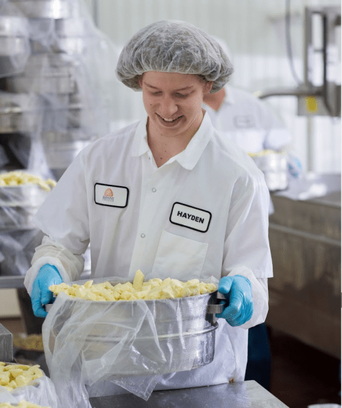 A worker in a hairnet and gloves smiles while carrying a large pot of chopped food in a kitchen setting.