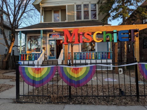 A colorful storefront with the word "Mischief" above the entrance, decorated with rainbow banners and an open sign.