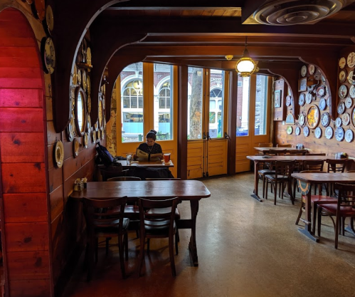A cozy restaurant interior with wooden walls, decorative plates, and a person reading at a table by the window.