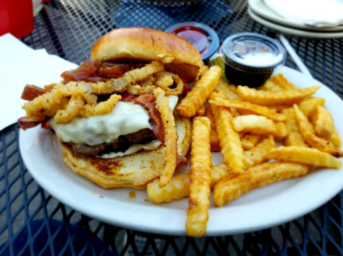 A hearty burger topped with bacon and onion rings, served with crinkle-cut fries and dipping sauces on a plate.