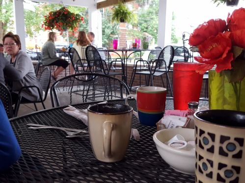 A cozy café scene with coffee cups on a table, surrounded by plants and patrons enjoying their time.