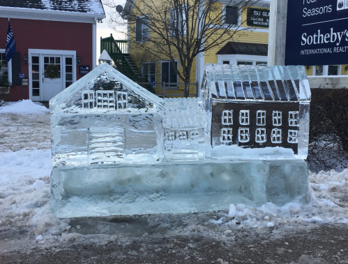 A clear ice sculpture of two buildings, one resembling a barn and the other a house, set in a snowy outdoor area.