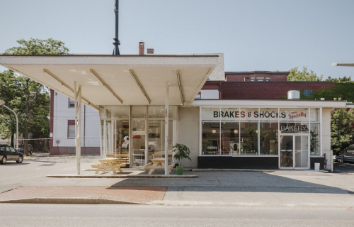 A modern bakery with outdoor seating, featuring large windows and a sign that reads "Brakes & Shocks Bakery."