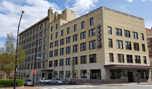 A large, brick building with "PRESS" signage, located at a street corner under a partly cloudy sky.