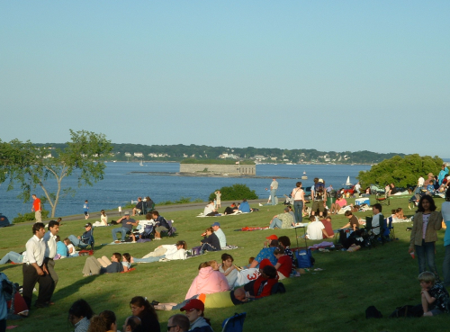 A crowded park by the water, with people lounging on the grass and a distant fort visible across the bay.