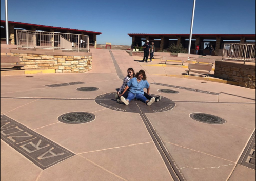 Two children sit on a compass rose design in a plaza, with buildings and people in the background under a clear blue sky.