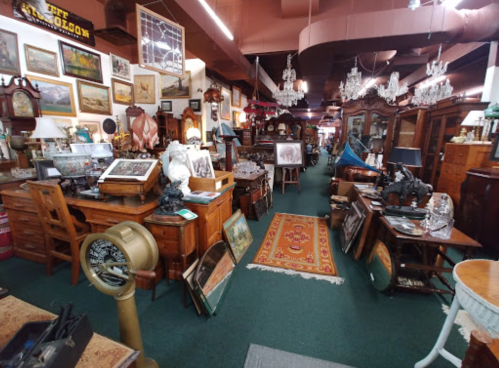 A vintage shop filled with antique furniture, artwork, and decor, featuring wooden tables, chandeliers, and a colorful rug.