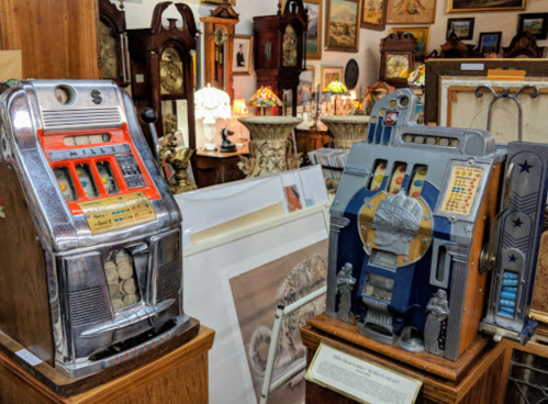 Two vintage slot machines displayed in an antique shop, surrounded by various artworks and decor.