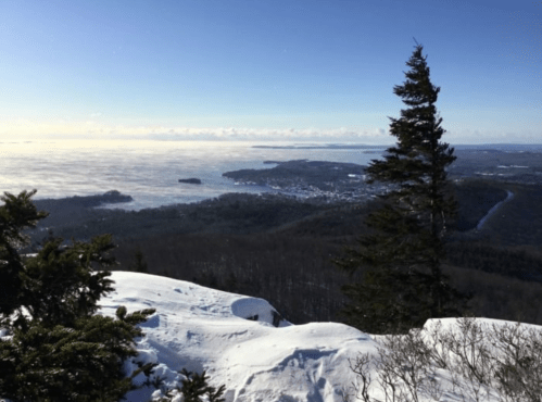 A snowy mountain landscape with a view of a distant lake and islands under a clear blue sky.