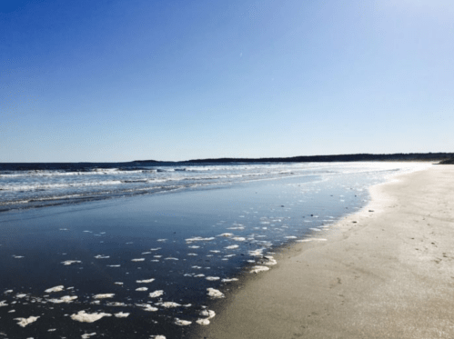 A serene beach scene with gentle waves, foam on the shore, and a clear blue sky.