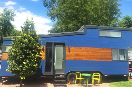 A blue and wood-paneled tiny house on wheels, surrounded by greenery and featuring a small outdoor seating area.