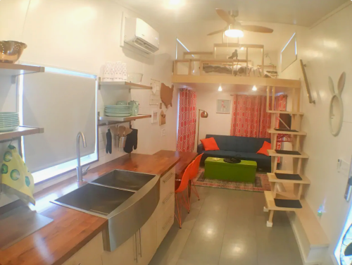 A modern tiny home interior featuring a kitchen, lofted bed, and cozy living area with a sofa and colorful decor.