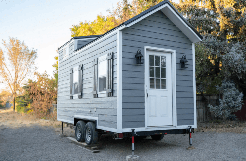 A small gray and white tiny house on wheels, featuring a front door and windows, set in a natural outdoor environment.
