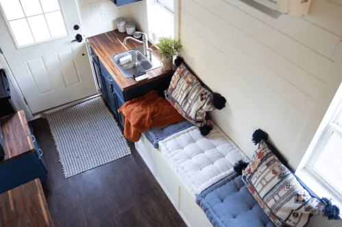 Cozy interior of a tiny home featuring a kitchen sink, wooden countertop, and cushioned seating by the window.
