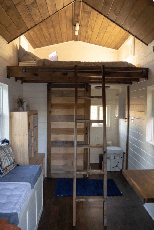 Cozy tiny home interior featuring a loft bed, wooden ladder, seating area, and compact bathroom.