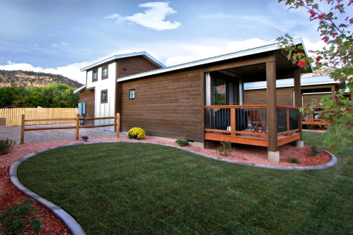 A modern wooden cabin with a porch, surrounded by green grass and landscaped gardens under a blue sky.