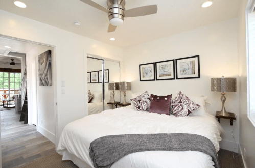 Cozy bedroom with a white bed, decorative pillows, wall art, and a ceiling fan, featuring natural light and modern decor.