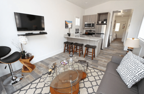 Modern living room with a TV, kitchen area, bar stools, and stylish decor, featuring a glass coffee table and cozy seating.
