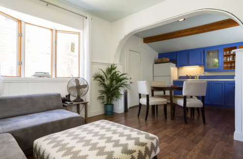 Cozy living space with a gray sofa, patterned ottoman, dining table, and blue kitchen cabinets in a bright room.